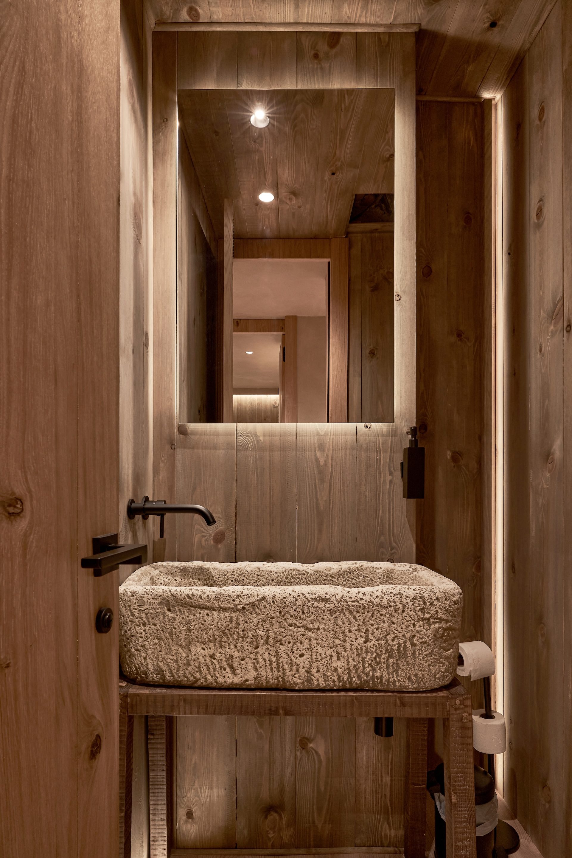 Lavabo de piedra tallada sobre mueble de madera en el aseo del Ático Nevado, con paredes de tablones y espejo retroiluminado