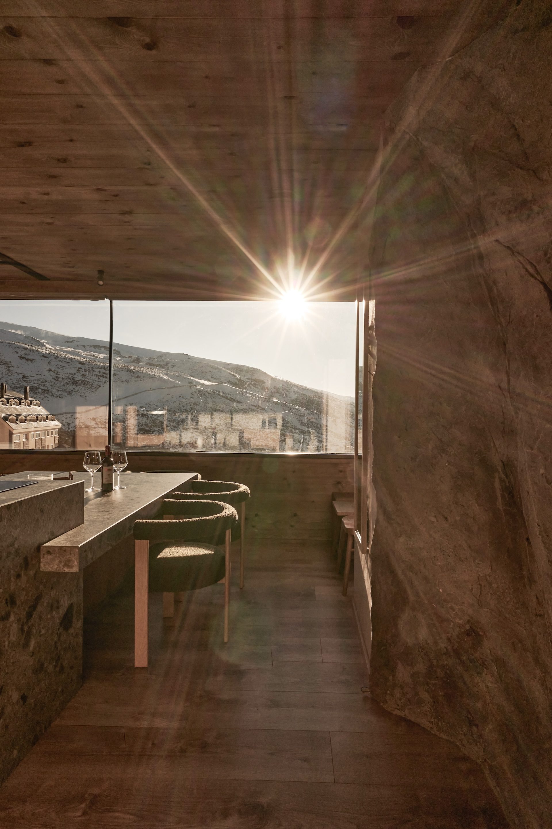 Comedor del Ático Nevado con sillas tapizadas, barra de piedra y el sol entrando por el ventanal sobre la sierra