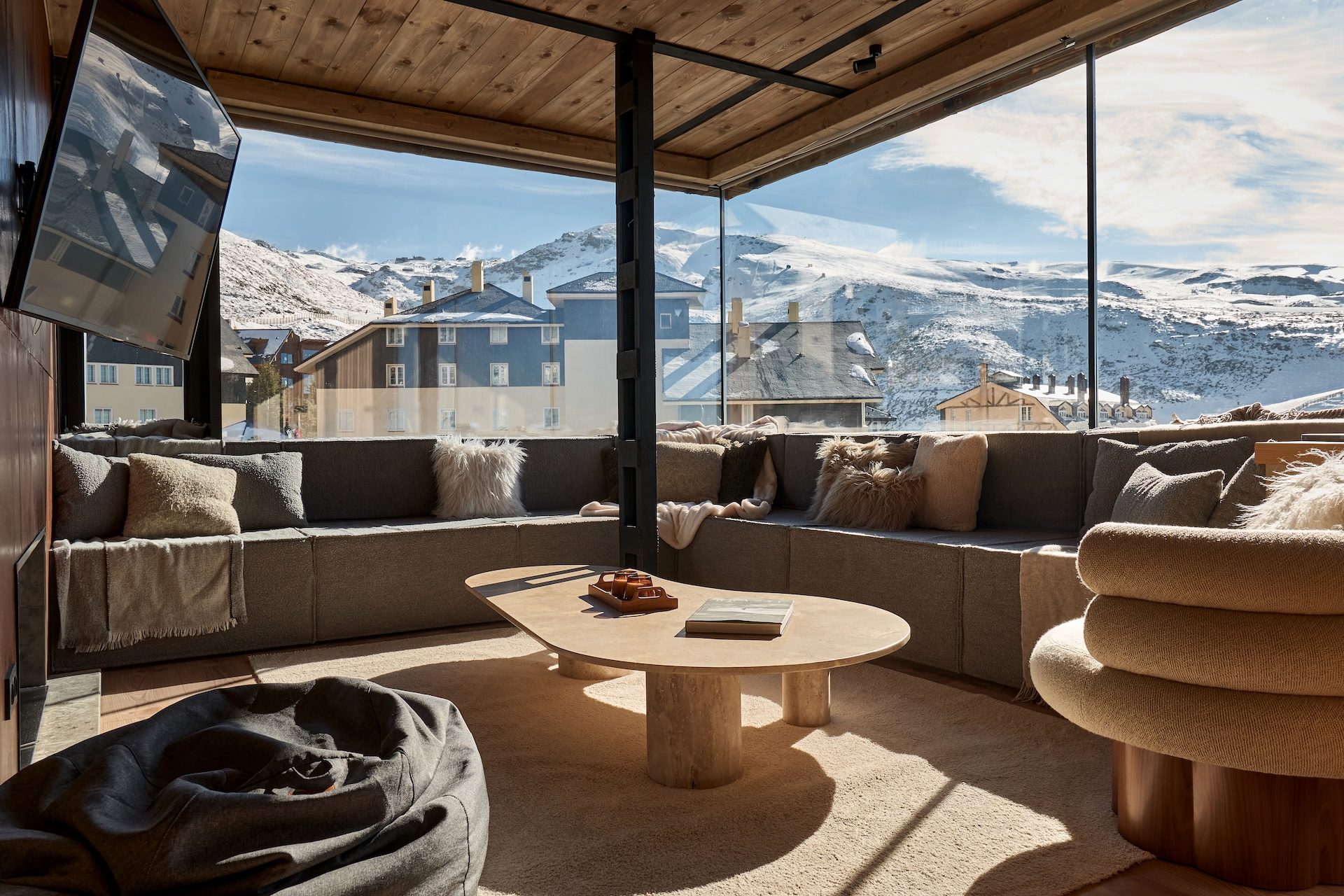 Salón de día del Ático Nevado: sofá corrido, mesa de travertino y ventanales abiertos a las montañas nevadas
