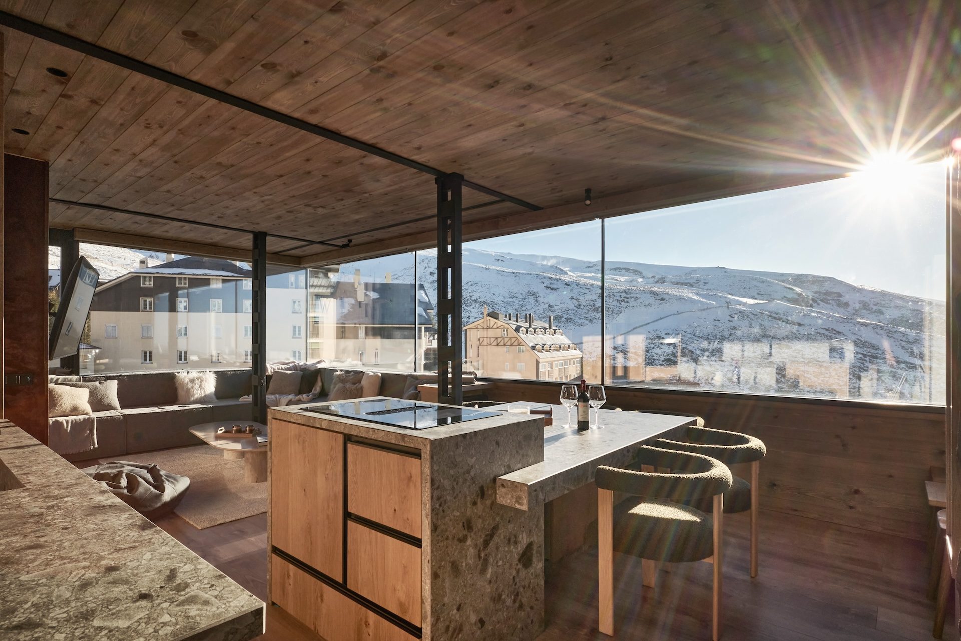 Cocina abierta del Ático Nevado con isla de piedra y madera, sillas tapizadas y el sol sobre las pistas de Sierra Nevada