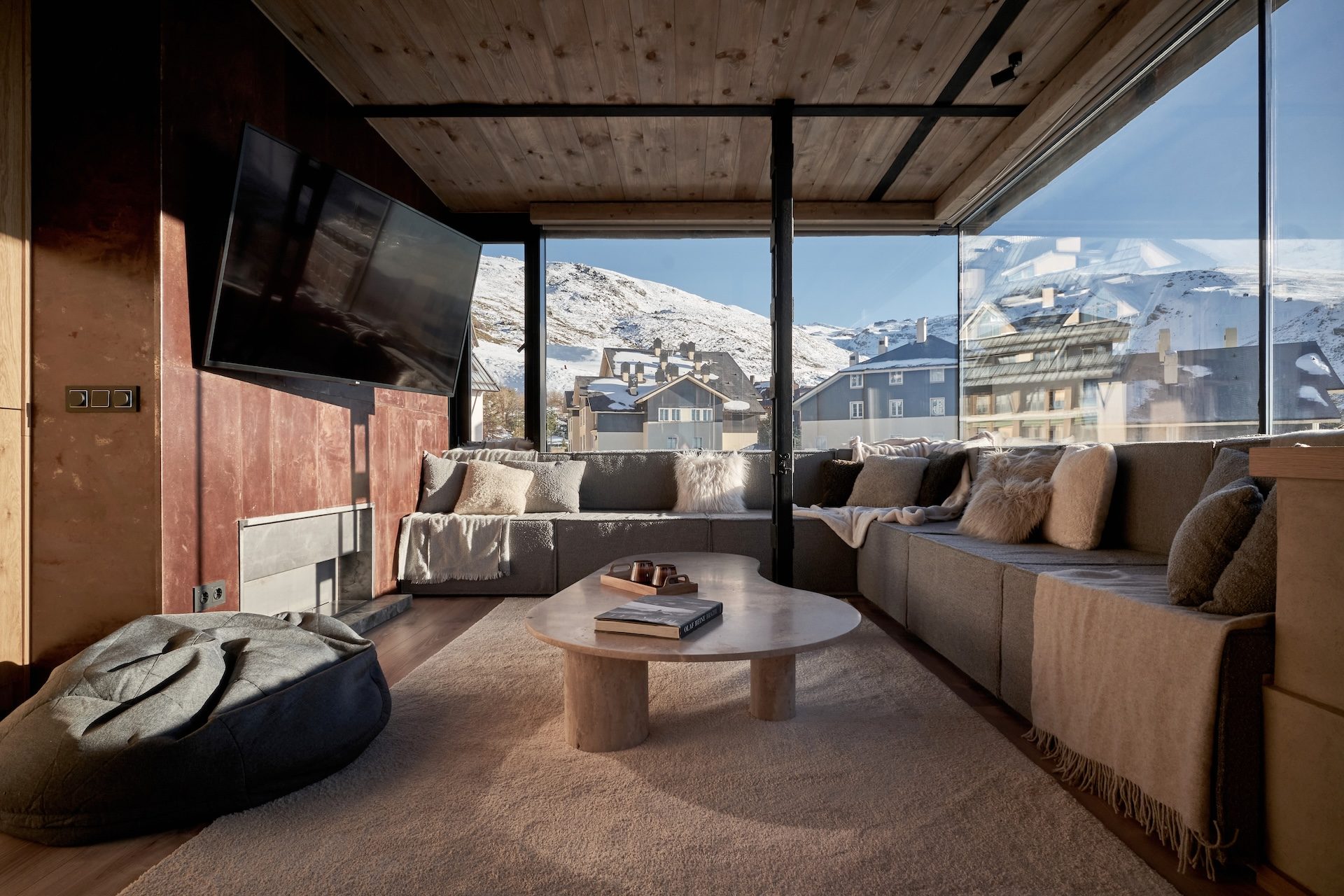 Salón del Ático Nevado en Sierra Nevada, Monachil: sofá corrido bajo techo de madera, mesa de travertino y grandes ventanales a las montañas nevadas