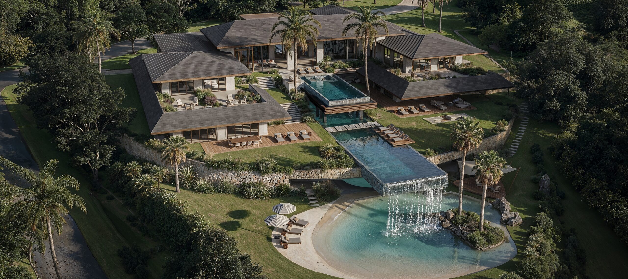 印度尼西亚热带豪华别墅建筑：Villa Sulawesi 鸟瞰，深色瓦屋顶、抬升泳池、瀑布与棕榈环绕的人工沙滩 · Archidom Studio